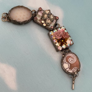 May include: A vintage bracelet featuring a series of ornate, silver-toned links. The bracelet incorporates various decorative elements, including a round stone, square panels with floral and crystal accents, and an oval charm with a swirl design. The bracelet is set against a light blue background.