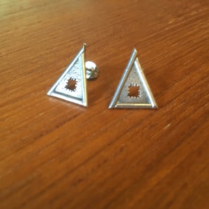 Könnte beinhalten: Ein Paar silberfarbene Dreiecks-Ohrstecker. Jeder Ohrring hat einen dreieckigen Rahmen mit einem quadratischen Ausschnitt in der Mitte. Die Ohrringe werden auf einer Holzoberfläche präsentiert.