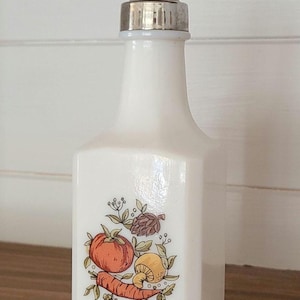 May include: A white glass cruet with a silver metal cap. The cruet features a colourful illustration of vegetables, including a tomato, carrot, and mushroom. The cruet is likely for holding oil or vinegar.