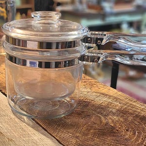 May include: A clear glass teapot with a lid and two handles. The teapot has a silver band around the middle and a clear glass lid. The handles are also clear glass. The teapot is sitting on a wooden surface.