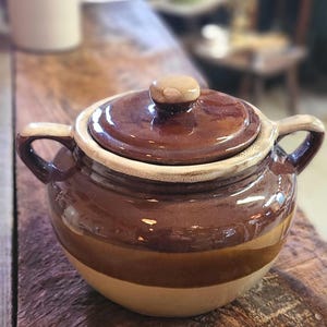 Peut inclure: Une marmite en céramique brune et beige avec un couvercle. La marmite a un corps large et rond et deux poignées. Le couvercle a un bouton sur le dessus.