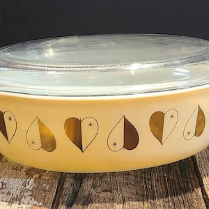 May include: An oval casserole dish with a clear glass lid. The cream-coloured dish features a pattern of gold hearts and starbursts. It has handles and is on a wooden surface. The dish is likely vintage.