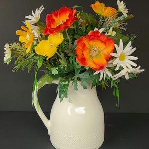 Peut inclure: Un pichet en céramique blanche avec un motif décoratif contient un bouquet de fleurs artificielles. Les fleurs sont un mélange de jaune, d'orange et de blanc, avec une variété de textures et de formes.