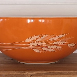 May include: Orange mixing bowl with a wheat stalk design. The bowl has a wide, rounded shape with a handle on each side. The wheat stalks are white and are arranged horizontally across the bowl.