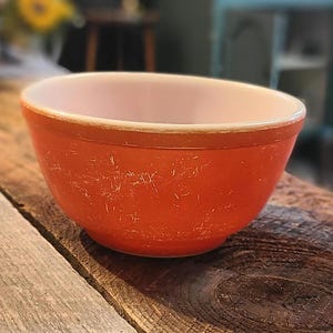 May include: A vintage orange mixing bowl with a cream interior. The bowl has a rounded shape and a slightly worn appearance. The bowl is on a wooden surface. The bowl's diameter is approximately 15 cm.