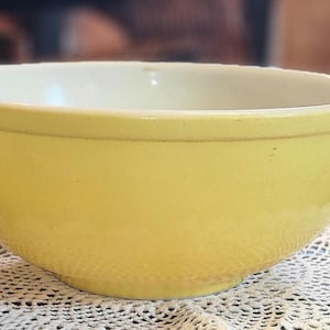 May include: A vintage yellow mixing bowl with a cream interior. The bowl has a rounded shape and a slightly flared rim. It is resting on a white lace doily.