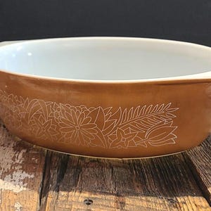 May include: A brown casserole dish with a white interior and handles. The exterior has a white floral design. This vintage dish is suitable for baking and serving.