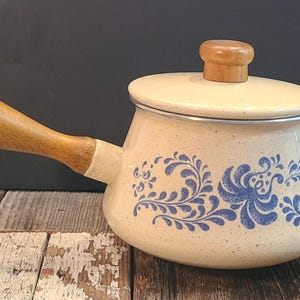 May include: Cream-coloured enamel saucepan with a wooden handle and lid. The pot features a blue floral design. The handle and lid knob are made of wood. The pot is on a wooden surface.