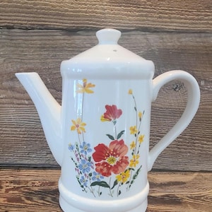 May include: White ceramic teapot with a lid and handle, decorated with a floral pattern of red, yellow, and blue flowers. The teapot has a spout and a decorative base. The background is a wooden surface.