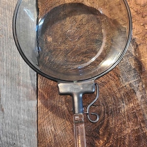 May include: A vintage glass and metal kitchen utensil, possibly a food mill or strainer. The clear glass bowl is held by a metal frame and handle. The handle is clear plastic. The utensil is set against a rustic wooden background.