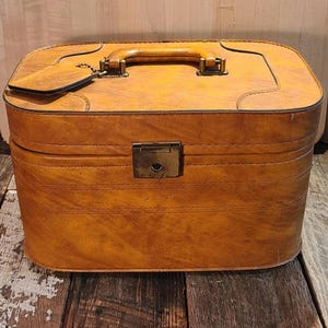 May include: A vintage, rectangular, tan suitcase with a leather handle and a metal clasp. The suitcase has a tag attached to the handle and is made of a textured material. The suitcase is sitting on a wooden surface.