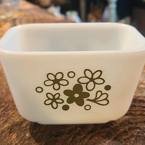May include: A square, white glass bowl with rounded corners. The bowl features an olive green floral design. The flowers are stylized and clustered on the front. Likely a vintage piece, the bowl is a kitchenware item.