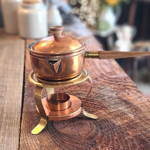 May include: A copper pot with a wooden handle sits on a brass stand with a small copper cup. The pot has a small spout and a knob on the lid.