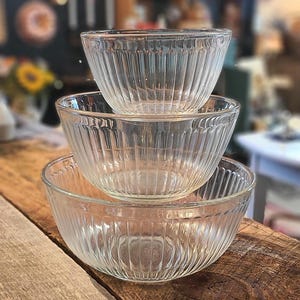 May include: Three clear glass mixing bowls stacked on top of each other. Each bowl features a ribbed design and a rounded shape. The bowls are nested, with the largest at the bottom and the smallest at the top. They are on a wooden surface.