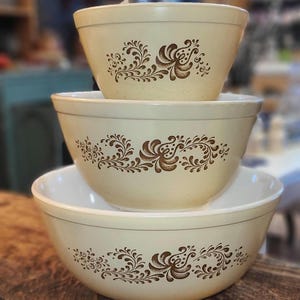 May include: Three nesting mixing bowls with a brown floral pattern on a beige background. The bowls are made of glass and are in various sizes.