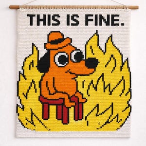 Könnte beinhalten: Ein gehäkeltes Wandbild mit dem Meme "This is Fine". Das Bild zeigt einen Hund mit orangefarbenem Hut, der auf einem roten Hocker sitzt und von gelben Flammen umgeben ist. Der Text "THIS IS FINE." steht über dem Hund.