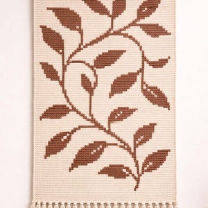 May include: Beige and brown woven wall hanging with a botanical leaf design. The leaves are a dark brown color and are arranged in a symmetrical pattern against a beige background. The wall hanging is finished with a fringe at the bottom and a wooden rod at the top.