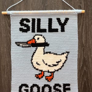 May include: Crocheted wall hanging with the words "SILLY GOOSE" in black block letters. A white goose with an orange beak and feet is holding a knife in its beak. The background is light blue, and the hanging is attached to a wooden dowel.