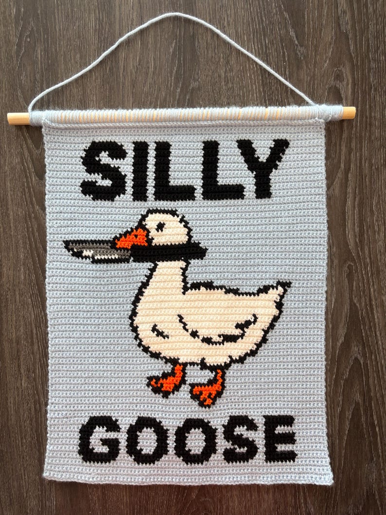 May include: Crocheted wall hanging with the words "SILLY GOOSE" in black block letters. A white goose with an orange beak and feet is holding a knife in its beak. The background is light blue, and the hanging is attached to a wooden dowel.
