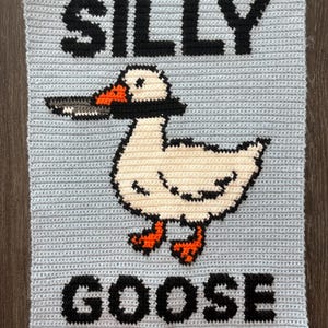 May include: A light blue crocheted square with the words "SILLY GOOSE" in black block letters. A cartoon goose with a knife in its beak is centered on the square. The goose is white with orange feet and beak accents.