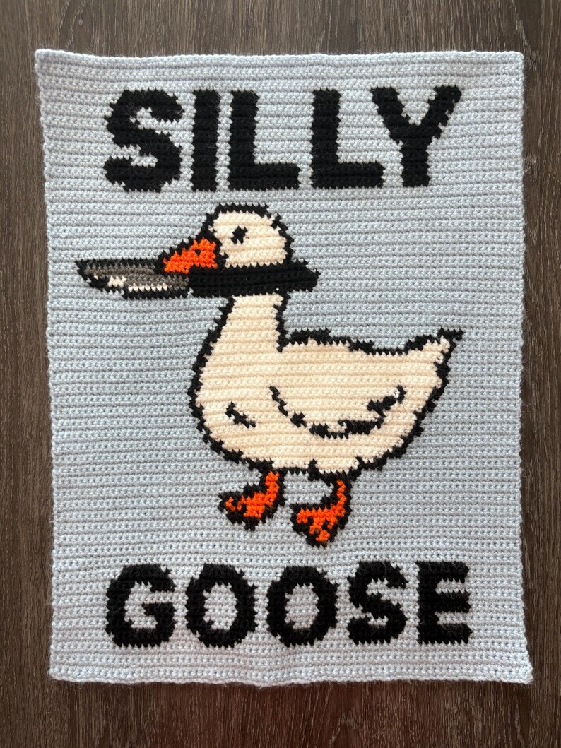 May include: A light blue crocheted square with the words "SILLY GOOSE" in black block letters. A cartoon goose with a knife in its beak is centered on the square. The goose is white with orange feet and beak accents.