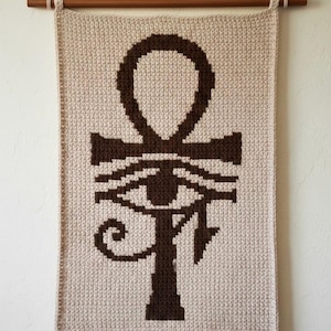 Pode incluir: Tapeçaria de parede de crochê bege com um símbolo Ankh e um design de Olho de Hórus em marrom escuro. A tapeçaria é suspensa por uma vara de madeira com um cordão de fibra natural. Decoração de parede única.