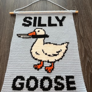 Peut inclure: Une tenture murale crochetée bleu clair avec les mots "SILLY GOOSE" en lettres noires. Un dessin d'oie avec un corps blanc, des pieds orange et un bec noir et gris est centré sous le texte. La tenture est fixée à une cheville en bois.