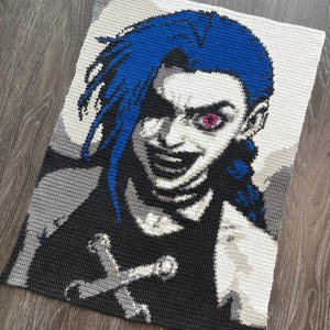 Puede incluir: Manta de ganchillo con un retrato estilizado de un personaje con cabello azul y una expresión siniestra. La obra de arte utiliza tonos de gris, negro, blanco y azul, con un toque de rosa en el ojo del personaje. El personaje lleva una camiseta negra con un diseño de calavera blanca.