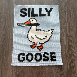 May include: A light blue crocheted square with the words "SILLY GOOSE" in black block letters. A white goose with an orange beak and feet is in the center, holding a knife in its beak. The goose is outlined in black.