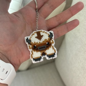 May include: A cross-stitch keychain featuring a cartoon character in brown, white, and gray. The keychain has a silver chain and ring. The character has a cross-stitch design and is in a standing pose. The keychain is a handmade item.