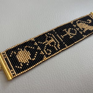 May include: A beaded bracelet featuring a black and gold pattern. The bracelet is rectangular with gold-colored metal end pieces and a delicate chain. The intricate design is made of small, evenly spaced beads.