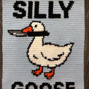 May include: A light blue, crocheted wall hanging with the words "SILLY GOOSE" in black block letters. A white goose with black accents and orange feet and beak is in the center, holding a cigarette. The wall hanging is mounted on a wooden dowel.