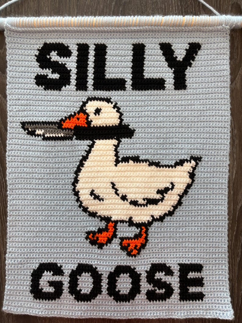 May include: A light blue, crocheted wall hanging with the words "SILLY GOOSE" in black block letters. A white goose with black accents and orange feet and beak is in the center, holding a cigarette. The wall hanging is mounted on a wooden dowel.