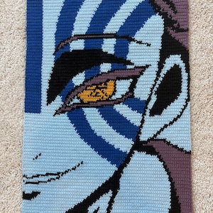 Può includere: Arazzo da parete all'uncinetto raffigurante un occhio stilizzato. L'opera d'arte utilizza filato blu, azzurro, nero, viola e giallo. L'occhio è il punto focale, con strisce concentriche blu e azzurre. Il pezzo è rettangolare.