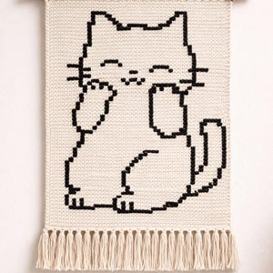 May include: A cream-colored macrame wall hanging featuring a black outline of a cat. The cat is depicted in a playful pose, with its paws near its face. The bottom edge has a fringe detail, and the piece is suspended from a wooden dowel.
