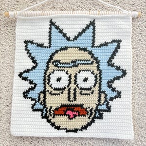 May include: White, crocheted wall hanging featuring a cartoon character's face with light blue hair and black outlines. The character has a surprised expression with an open mouth and pink tongue. A wooden dowel and white cord are at the top.