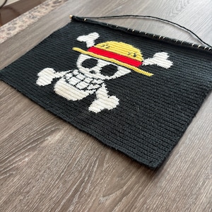 Może przedstawiać: Czarny obraz na ścianę z białym wzorem czaszki i skrzyżowanych piszczeli, zwieńczony żółtym i czerwonym kapeluszem. Wzór jest wyśrodkowany na czarnym tle. Do zawieszenia przymocowany jest drewniany pręt i czarny sznurek.