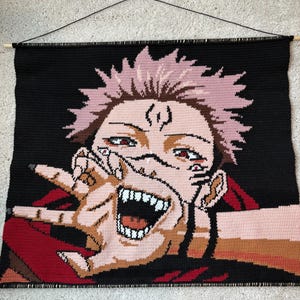 May include: A handmade, crocheted wall hanging depicting a character with pink hair and red eyes, set against a black backdrop. The artwork shows the character with an open mouth and is ready to hang with a wooden dowel. The piece is handmade.