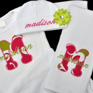 May include: A white baby outfit set with a hat, bodysuit, and burp cloth. The set is embroidered with the name "madison" in pink and green. The hat has a green flower with a rhinestone center.