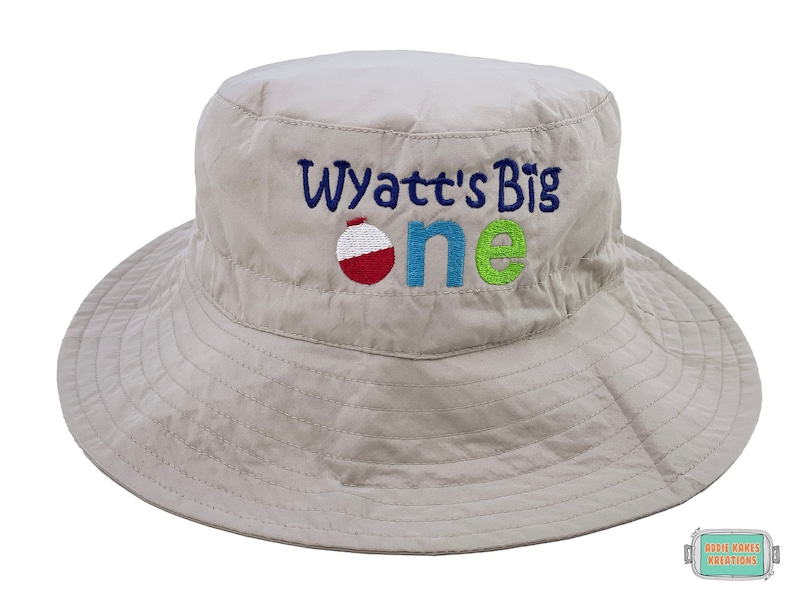 May include: A beige bucket hat with the text "Wyatt's Big One" embroidered on the front. The text is in blue, green, and red with a red and white fishing bobber in between the words "Big" and "One".