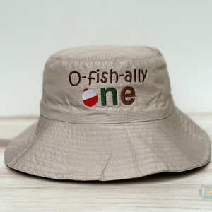 May include: A beige bucket hat with the text "O-fish-ally one" embroidered on the front. A red and white fishing bobber is embroidered next to the text.