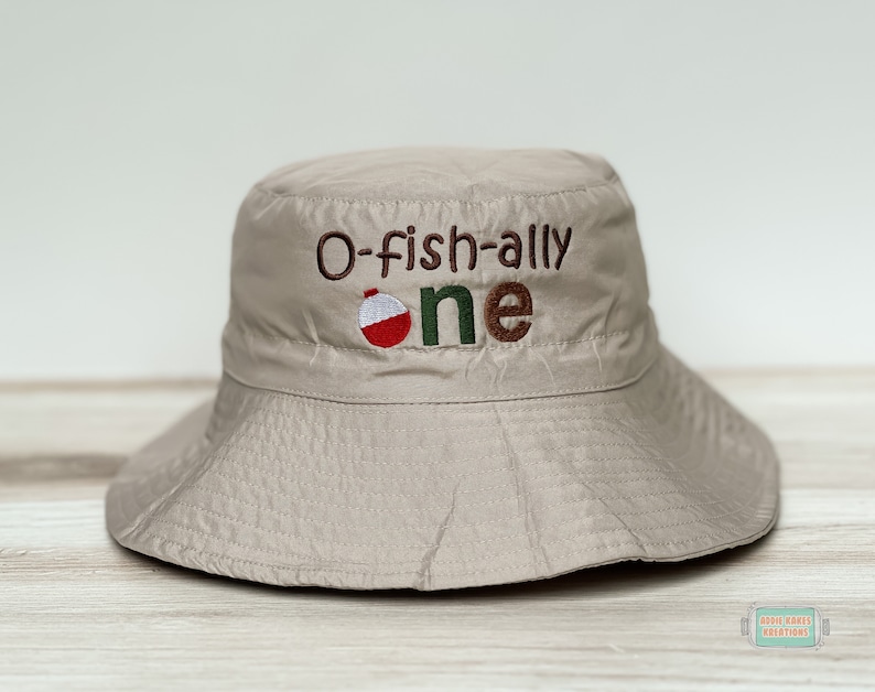 May include: A beige bucket hat with the text "O-fish-ally one" embroidered on the front. A red and white fishing bobber is embroidered next to the text.