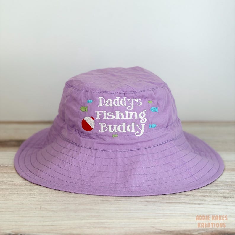 May include: A lavender sun hat with a wide brim. The hat features the embroidered text "Daddy's Fishing Buddy" in white, along with small embroidered fish and a red and white bobber. The hat is designed for sun protection.