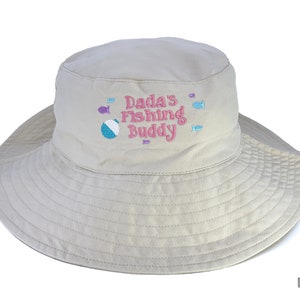 May include: A beige sun hat with a wide brim, featuring the embroidered text "Dada's Fishing Buddy" in pink, along with fish and a beach ball. The hat is designed for sun protection and outdoor activities.