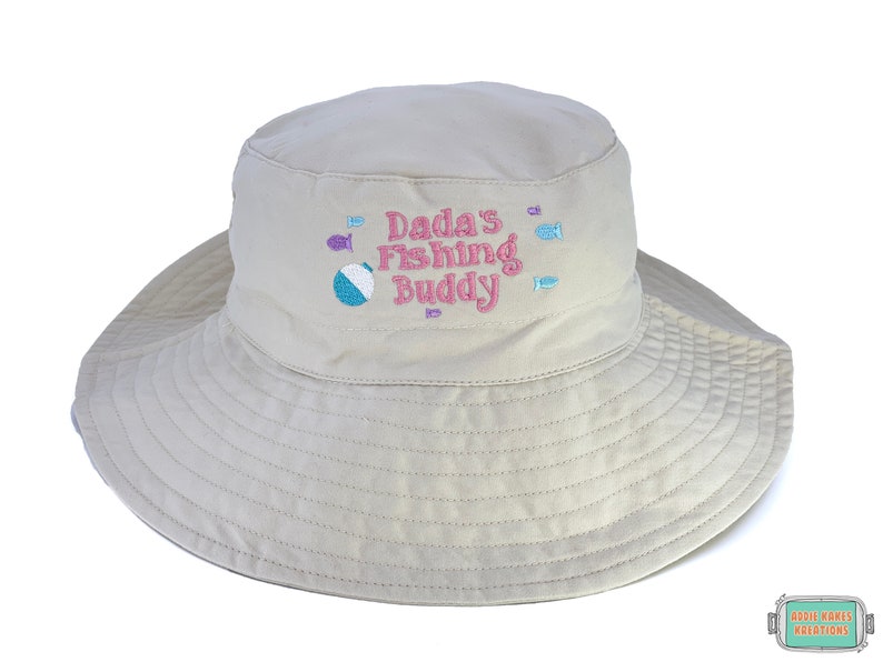 May include: A beige sun hat with a wide brim, featuring the embroidered text "Dada's Fishing Buddy" in pink, along with fish and a beach ball. The hat is designed for sun protection and outdoor activities.