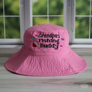 May include: A pink bucket hat with a wide brim. The hat features the embroidered text "Grandpa's Fishing Buddy" in black, along with small embroidered fish and a red and white bobber. The hat is made of a textured fabric.