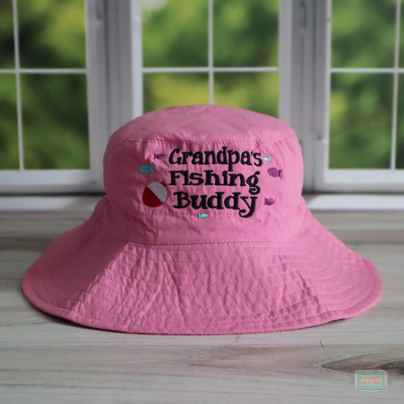 May include: A pink bucket hat with a wide brim. The hat features the embroidered text "Grandpa's Fishing Buddy" in black, along with small embroidered fish and a red and white bobber. The hat is made of a textured fabric.