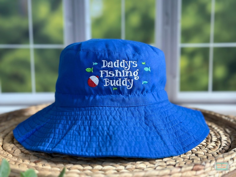 May include: A bright blue bucket hat with the embroidered text "Daddy's Fishing Buddy" in white. The hat features a red, white, and blue beach ball and small fish designs. The hat is sitting on a woven surface.