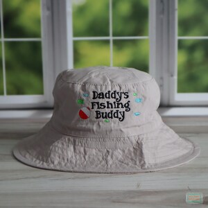 May include: A beige bucket hat with the embroidered text "Daddy's Fishing Buddy" in black, with colorful fish and a red and white bobber. The hat has a wide brim and is made of a soft fabric.