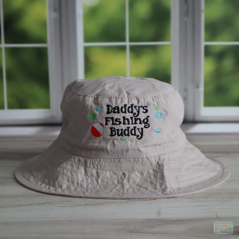 May include: A beige bucket hat with the embroidered text "Daddy's Fishing Buddy" in black, with colorful fish and a red and white bobber. The hat has a wide brim and is made of a soft fabric.